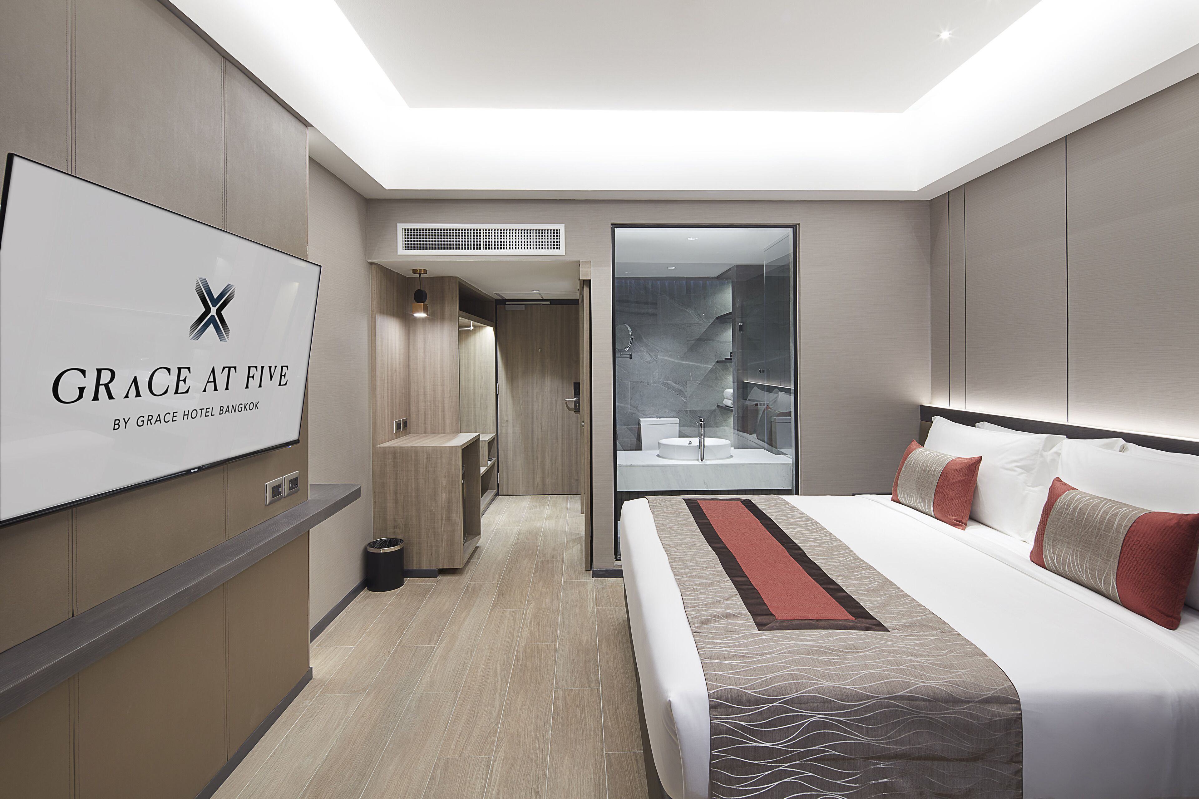 Ein modernes Hotelzimmer mit einem großen Bett, einem Fernseher, der den Namen des Hotels zeigt, einem Badezimmer mit Duschkabine und einer kleinen Sitzecke.
