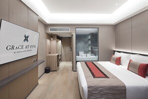 A modern hotel room with a large bed, a TV displaying the hotel's name, a bathroom with a glass shower, and a small seating area. - Grace At Five By Grace Hotel (Bangkok)