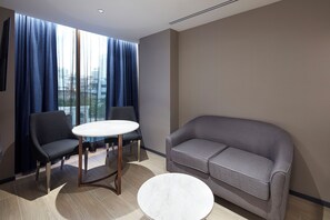 Ein modernes Hotelzimmer mit einer Couch, zwei Sessel, einem kleinen runden Tisch und großen Fenstern mit Vorhängen.