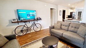 A living room with a grey sofa, a mounted TV, a bicycle, and a clock on the wall. - Rogers Retreat, Dog Friendly, Cozy Home. Close to Walmart Amp, Downtown Rogers (Rogers)