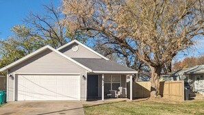 A single-story house with a garage, a covered porch, and a large tree in the background. - Rogers Retreat, Dog Friendly, Cozy Home. Close to Walmart Amp, Downtown Rogers (Rogers)