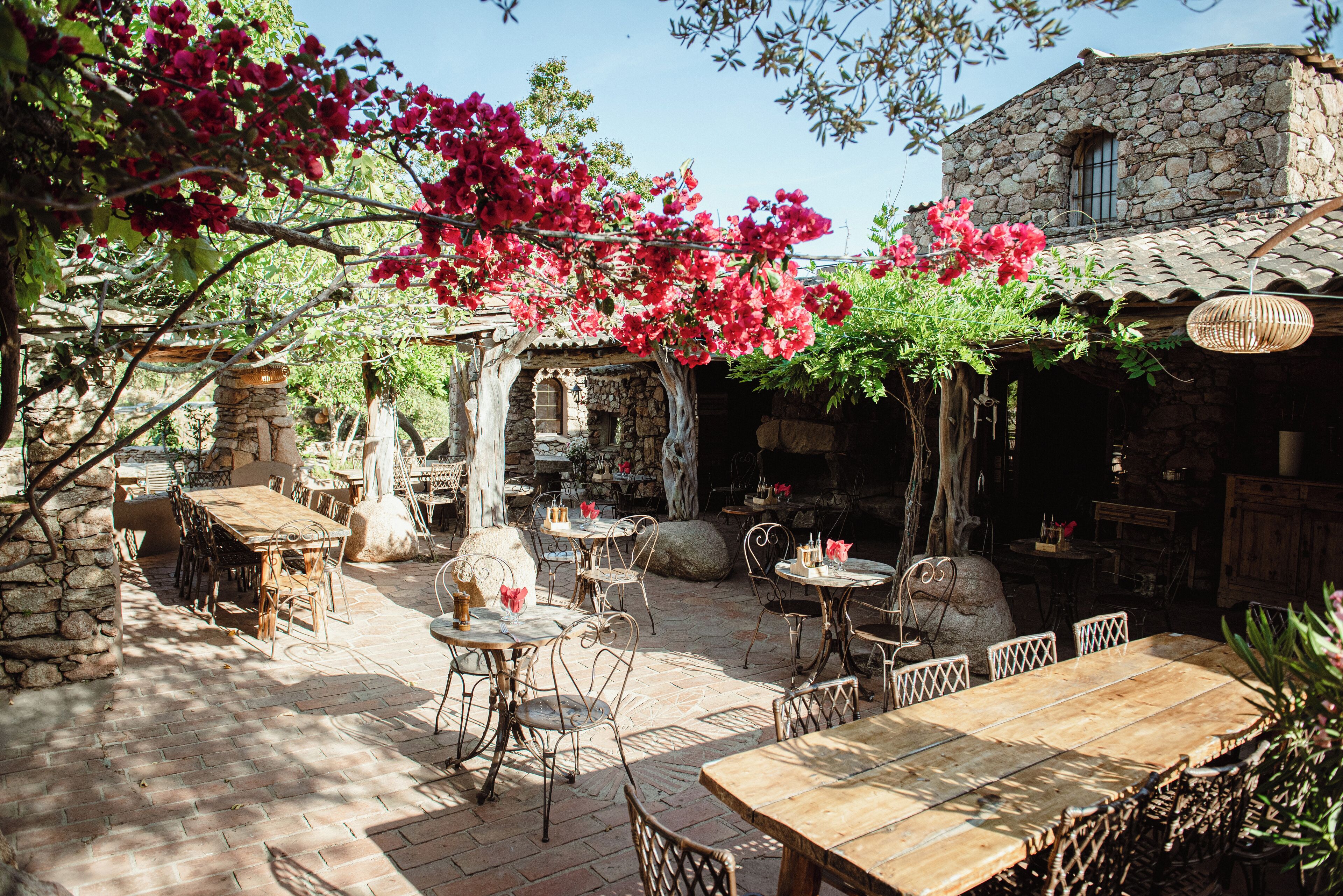 An outdoor patio area with stone walls, wooden tables and chairs, and a stone building in the background.