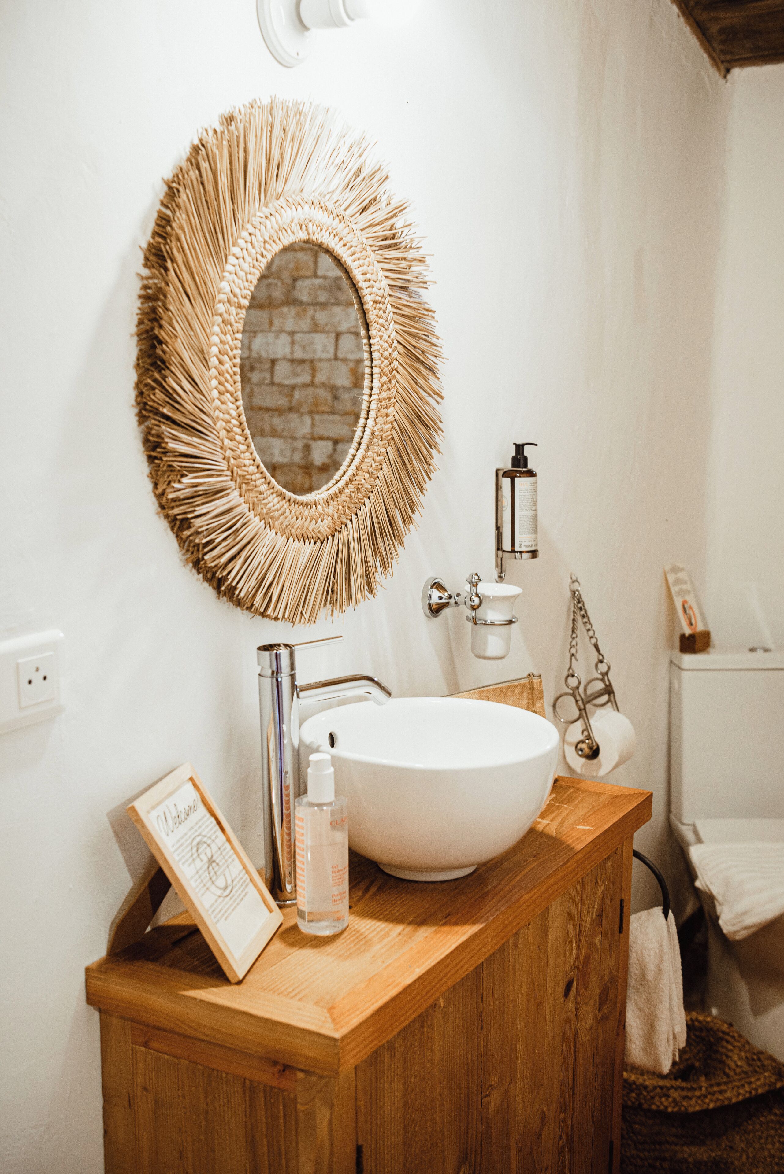 A bathroom with a wooden vanity, a white sink, a round mirror with a fringe design, and a brick wall behind the mirror.
