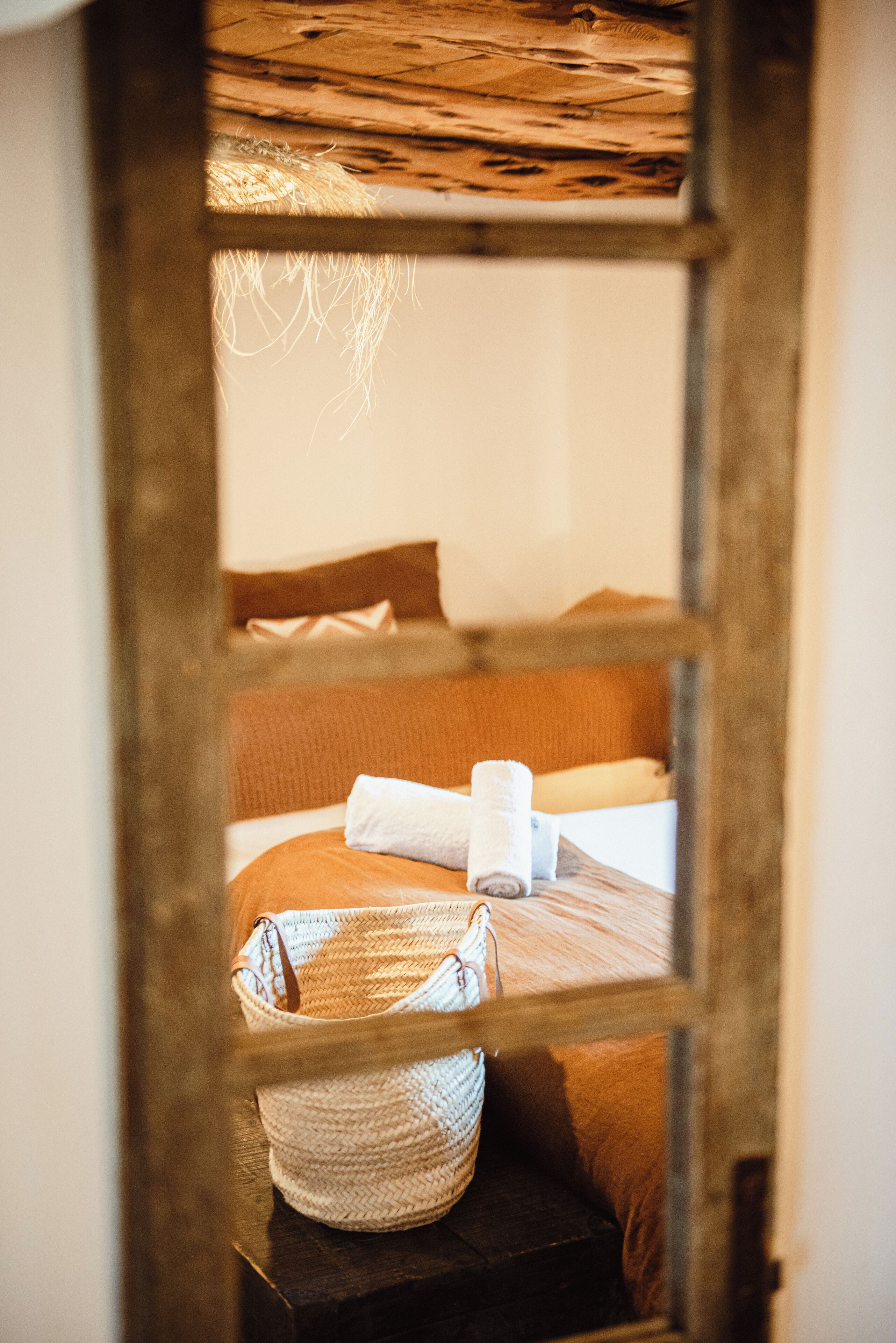 A rustic wooden-framed window with a bed visible through it, featuring a rolled towel and a woven basket.