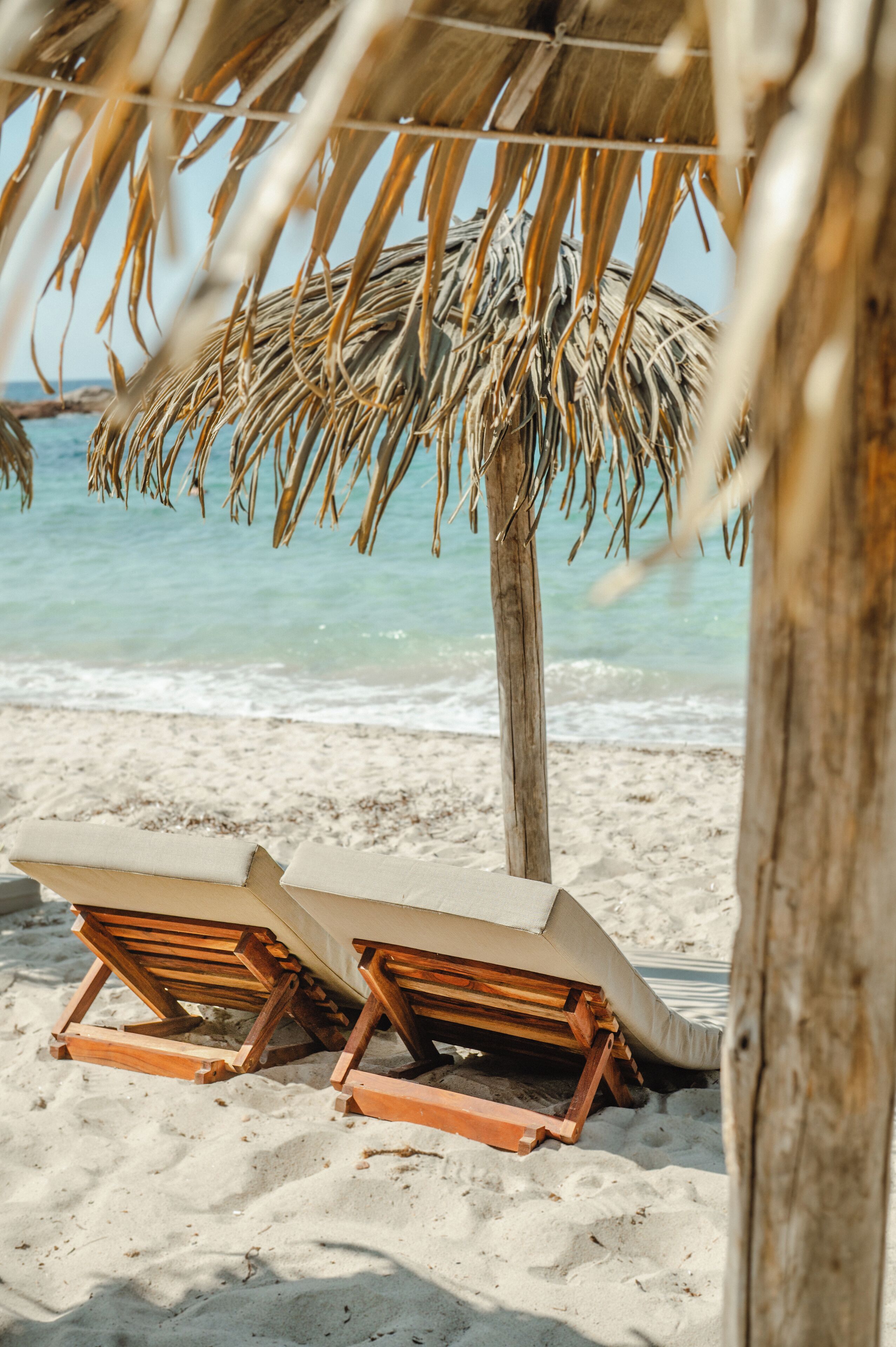 Twee houten lounge stoelen onder een rieten parasol op een zandstrand, met de oceaan op de achtergrond.