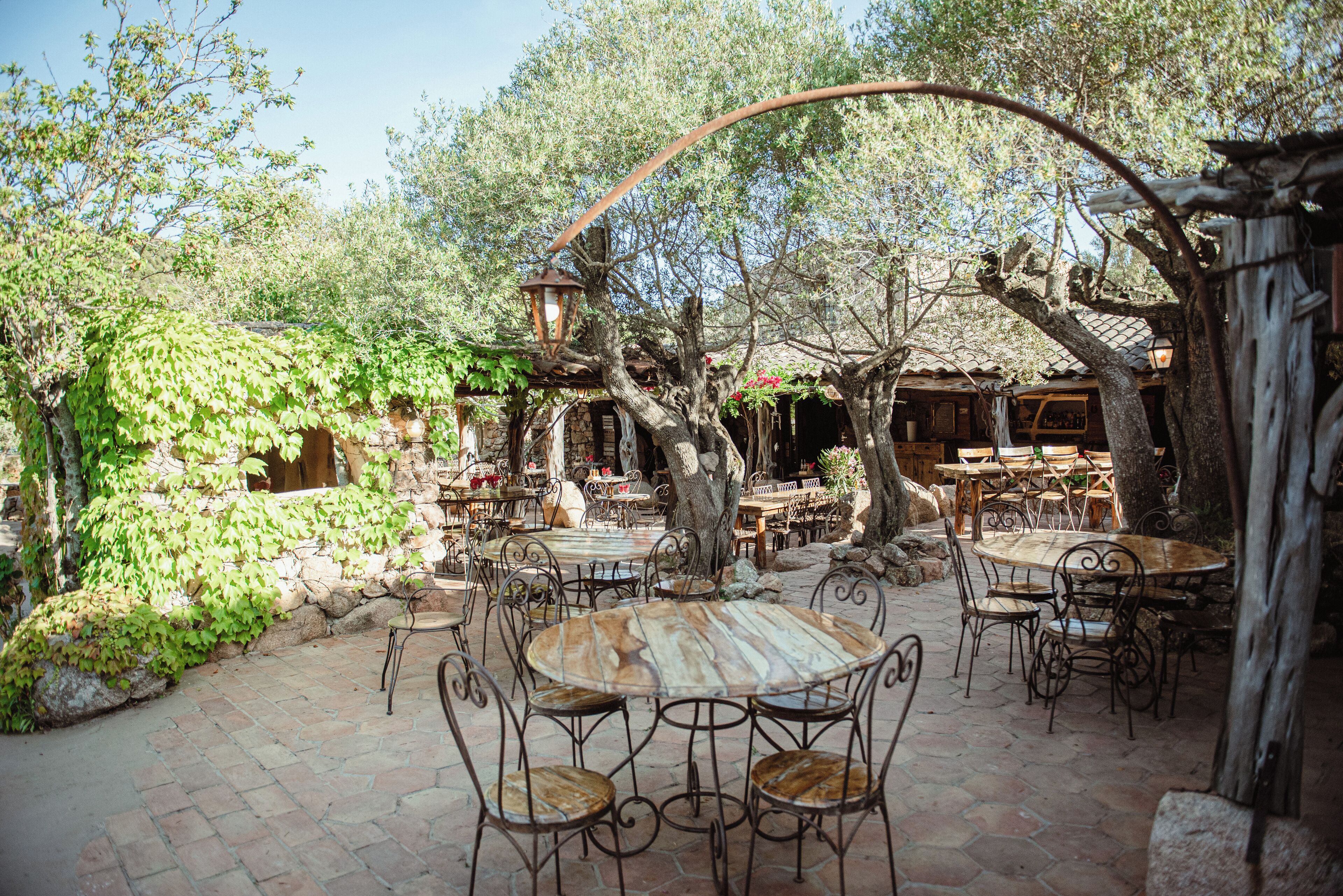 Une terrasse extérieure avec des tables et des chaises rondes, entourée d’arbres et de murs en pierre.