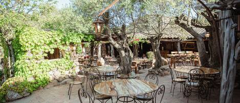 An outdoor patio with round tables and chairs, surrounded by trees and stone walls.