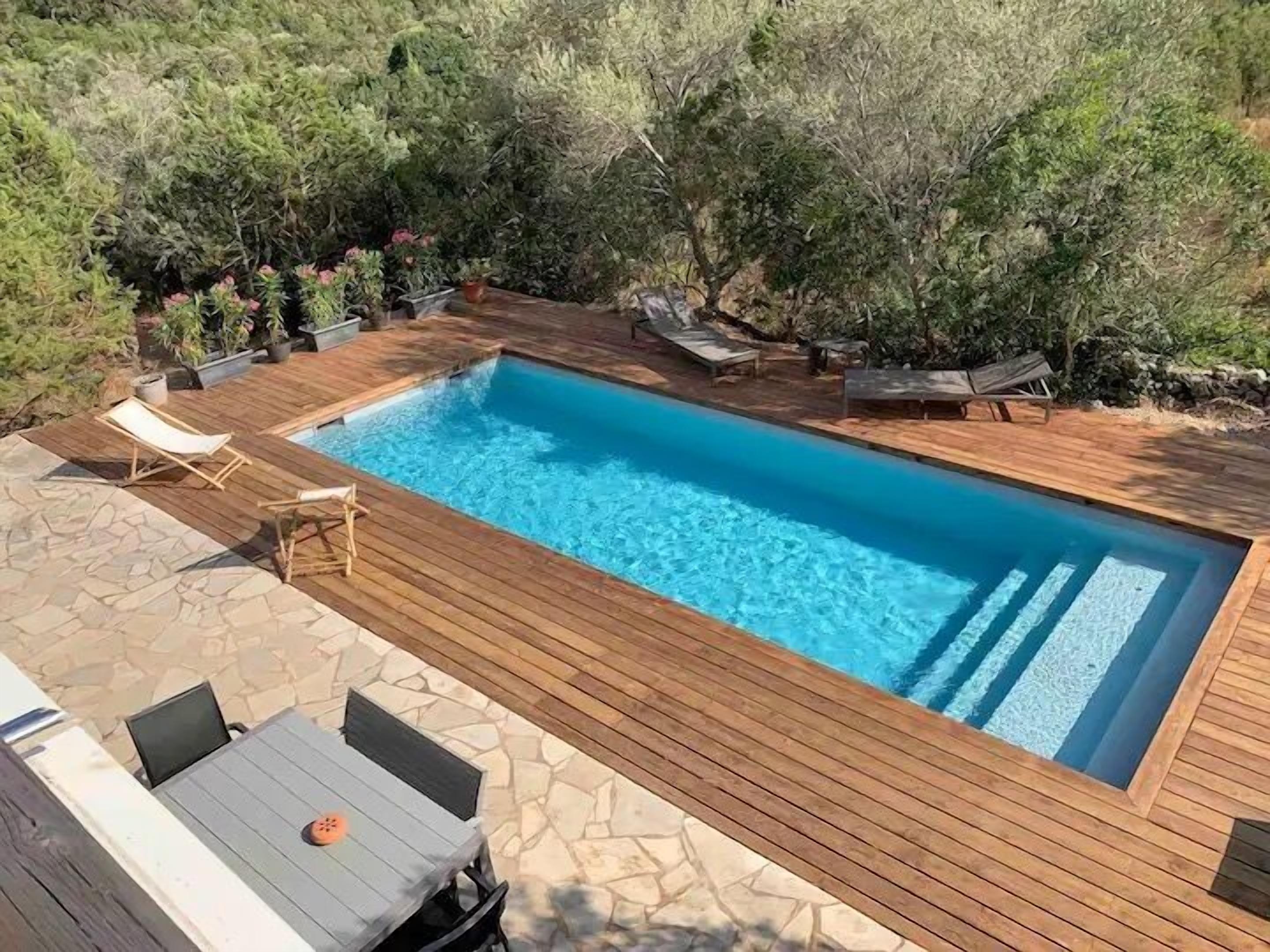 A rectangular swimming pool with a wooden deck, surrounded by lounge chairs and potted plants.