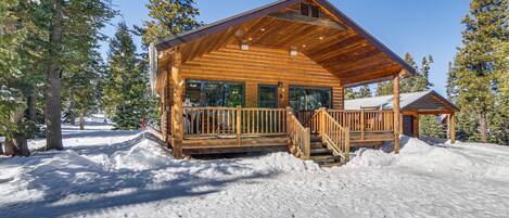 A wooden cabin with a covered porch and a staircase, surrounded by snow and trees.