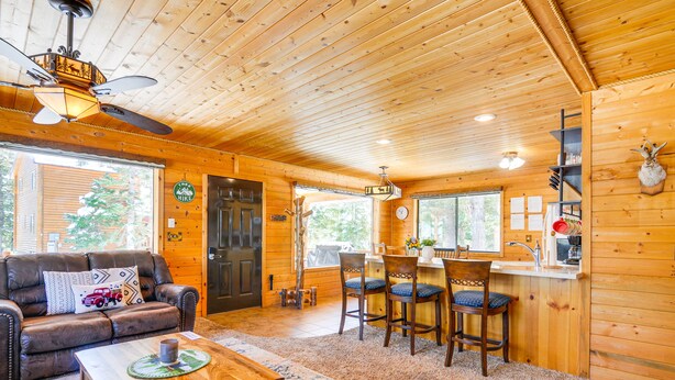 Een gezellig houten interieur met een houten plafond, een eethoek met tafel en stoelen, een leren bank met decoratief kussen, en een groot raam met uitzicht op bomen.