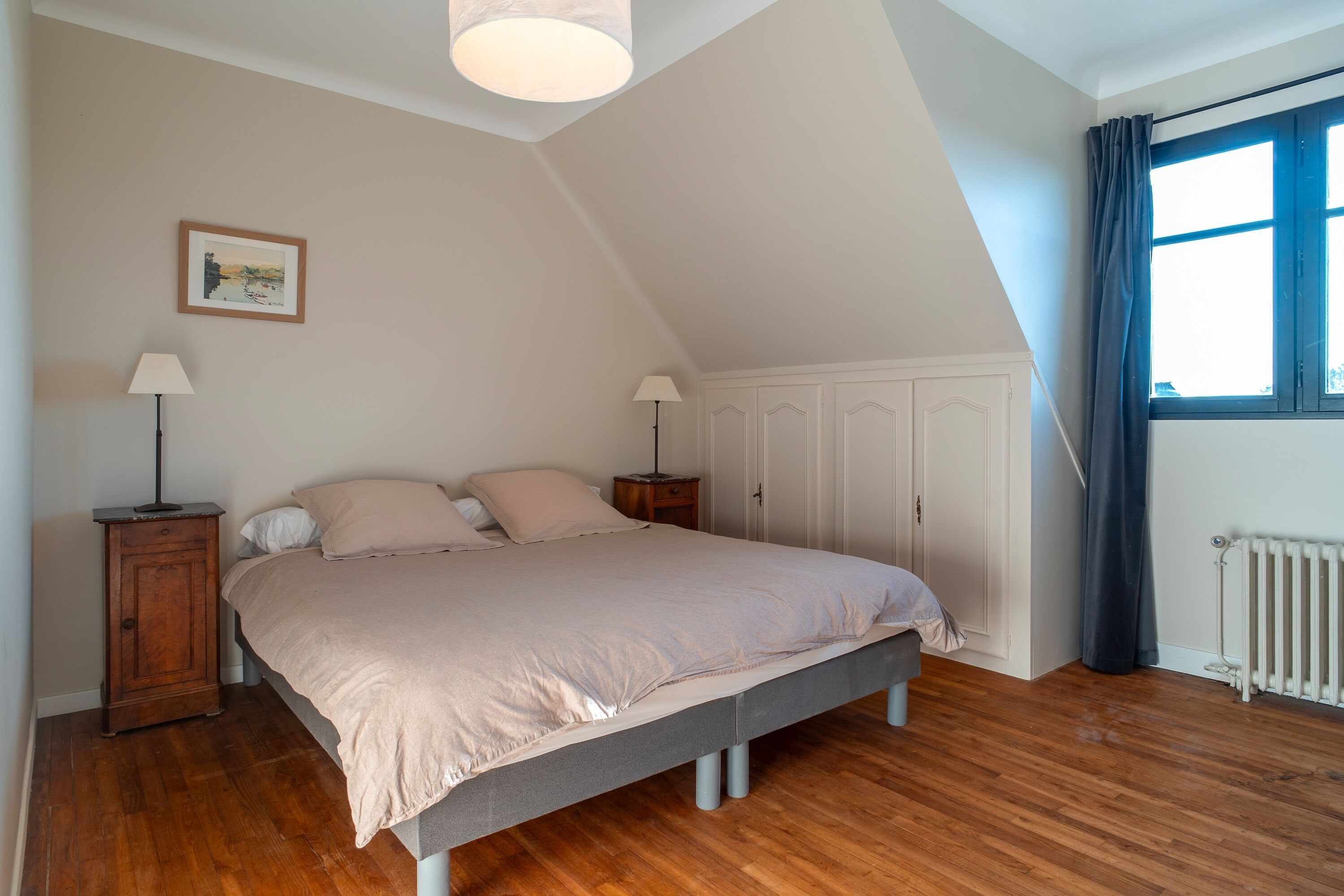 Une chambre à coucher avec un lit, deux tables de chevet, une lampe, un radiateur, une fenêtre avec des rideaux et un tableau au mur.