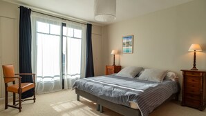 Un dormitorio con cama, una silla, una mesita de noche, una lámpara y un ventanal con cortinas.