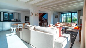 Amplia sala de estar con sofá, mesa de comedor y sillas, una cocina al fondo y una escalera que lleva a un nivel superior.