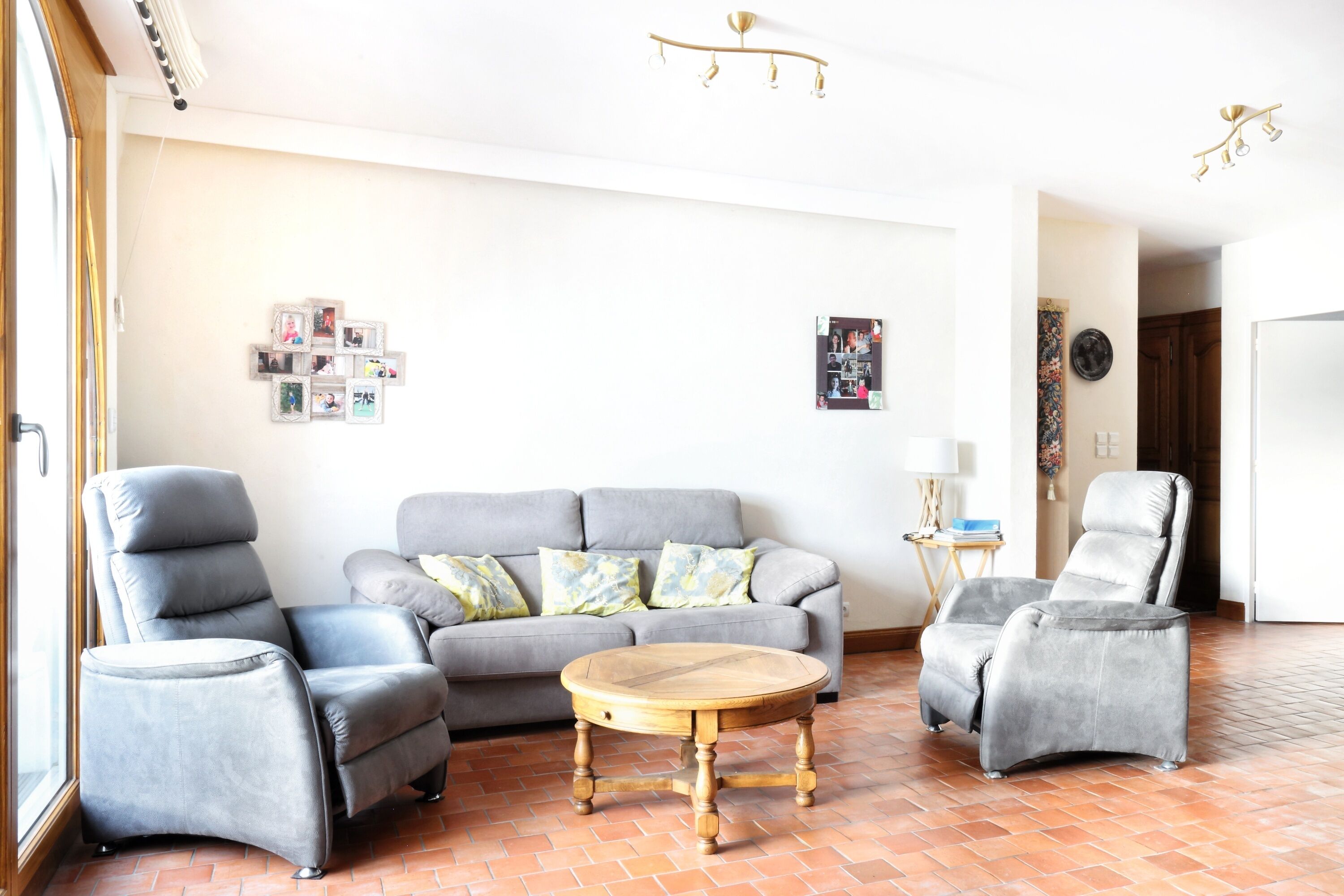 A living room with a grey sofa, two grey armchairs, a wooden coffee table, and a tiled floor.