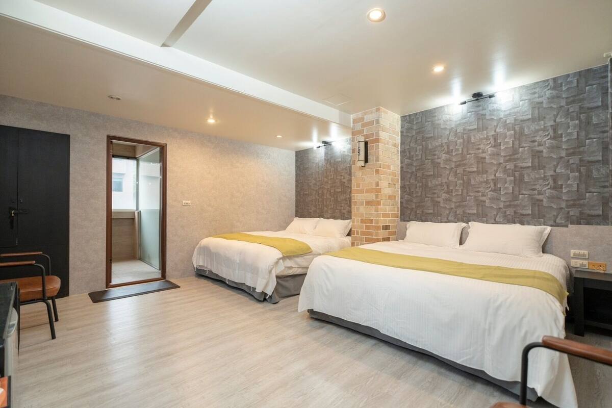 A hotel room with two beds, a desk, and a chair. There is a brick accent wall and a wooden floor.