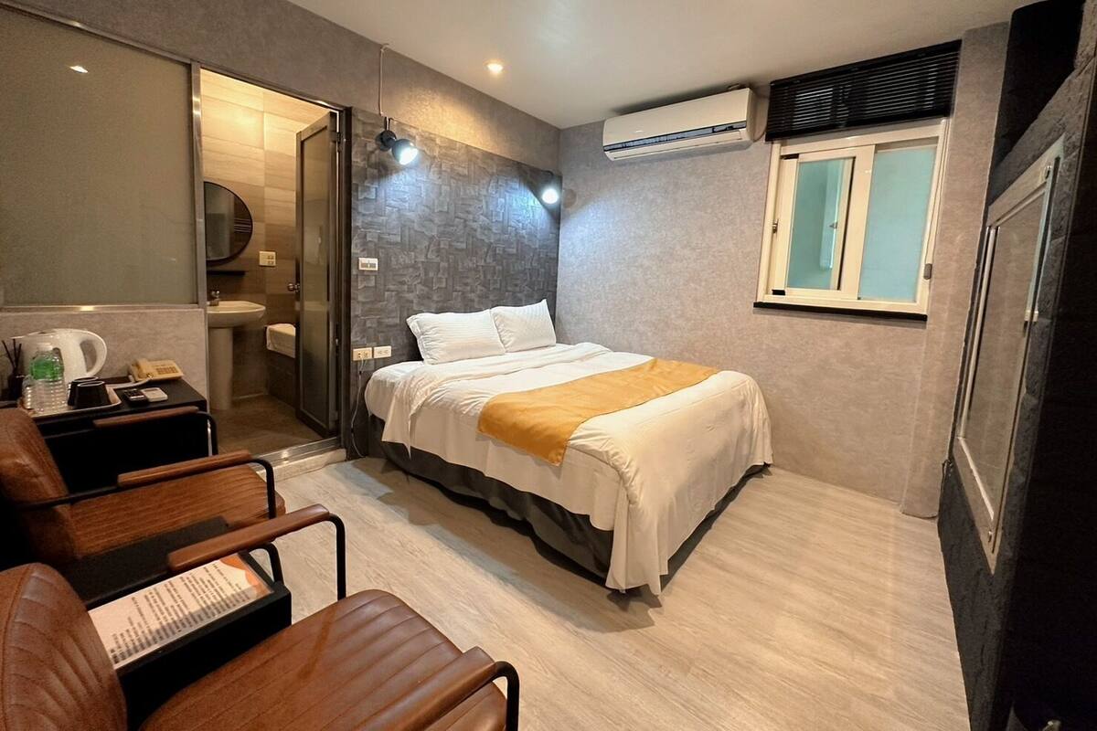 A modern hotel room with a bed, a desk, a chair, a bathroom, and an air conditioning unit.