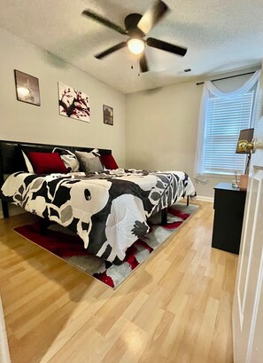 Un dormitorio con cama, ventilador de techo, mesita de noche y ventana con persianas.