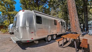 Un camping-car argenté est garé dans une zone boisée, à proximité d’une table de pique-nique et de quelques bancs.