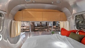 Interieur van een camper met een bed, een raam met gordijnen en uitzicht op bomen.
