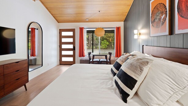 Un dormitorio con una cama grande, techo de madera, televisor, cómoda y una ventana con cortinas.