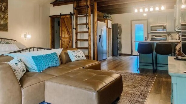 A spacious living area with a large brown leather sofa, a wooden staircase, and a kitchen area with modern appliances. - Enzo's Bungalow (Russellville)