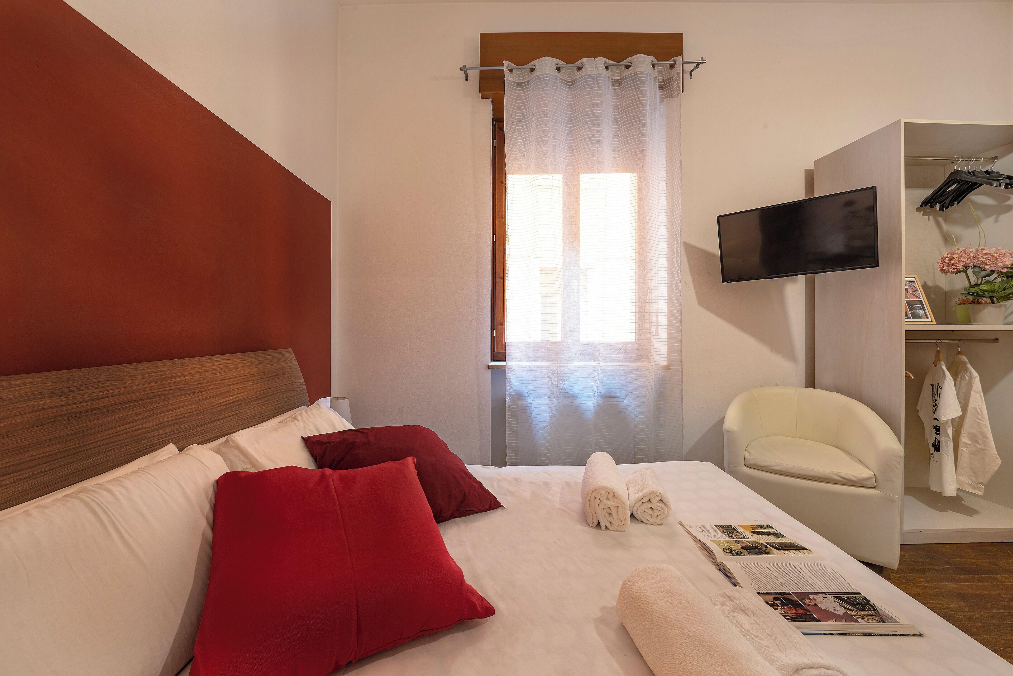 Quarto de hotel com cama, almofadas vermelhas, uma cadeira branca, televisão e janela com cortinas.