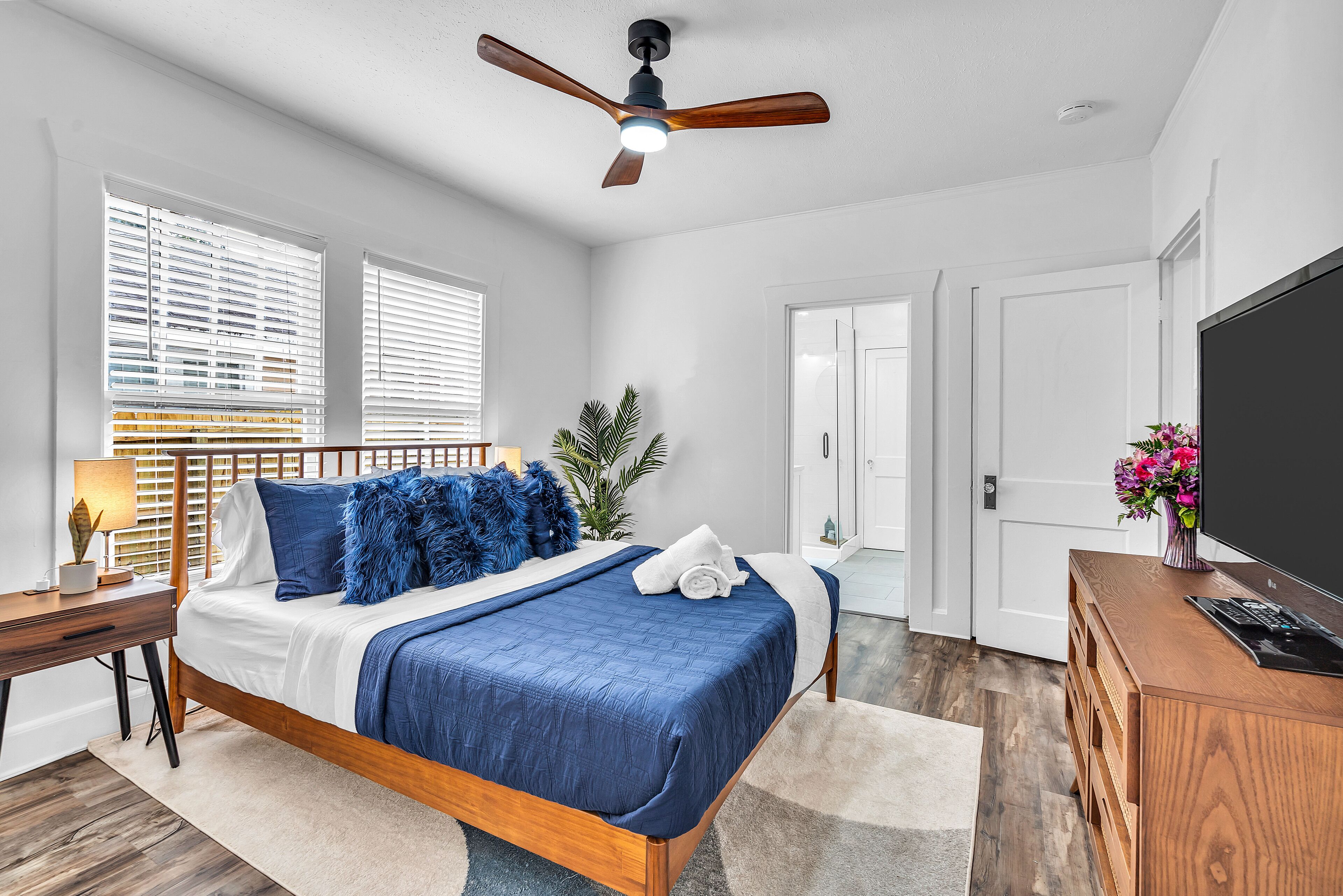 A bedroom with a bed, wooden headboard, ceiling fan, bedside table, lamp, and a TV on a wooden cabinet.