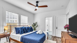A bedroom with a bed, wooden headboard, ceiling fan, bedside table, lamp, and a TV on a wooden cabinet.