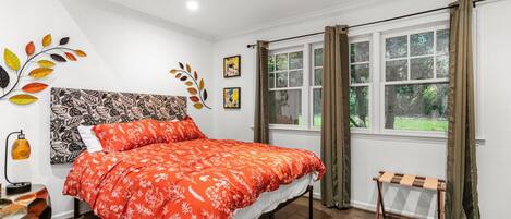 A bedroom with a bed, a bedside table, a lamp, a window with curtains, and a view of greenery outside.