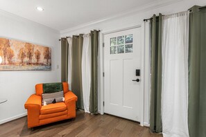 Une pièce avec une porte blanche, des rideaux verts, un plancher en bois et une chaise orange avec un coussin.