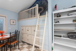 Une pièce avec un lit en mezzanine, un coin repas avec une table et des chaises, une étagère blanche où sont pliés des serviettes, et un plafond à motifs.
