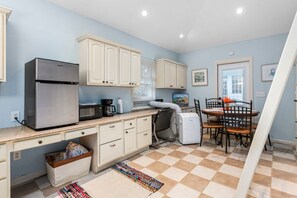 Eine kompakte Küche mit Kühlschrank, Mikrowelle und Kaffeemaschine. Ein Esstisch mit Stühlen und ein Teppichboden sind vorhanden.