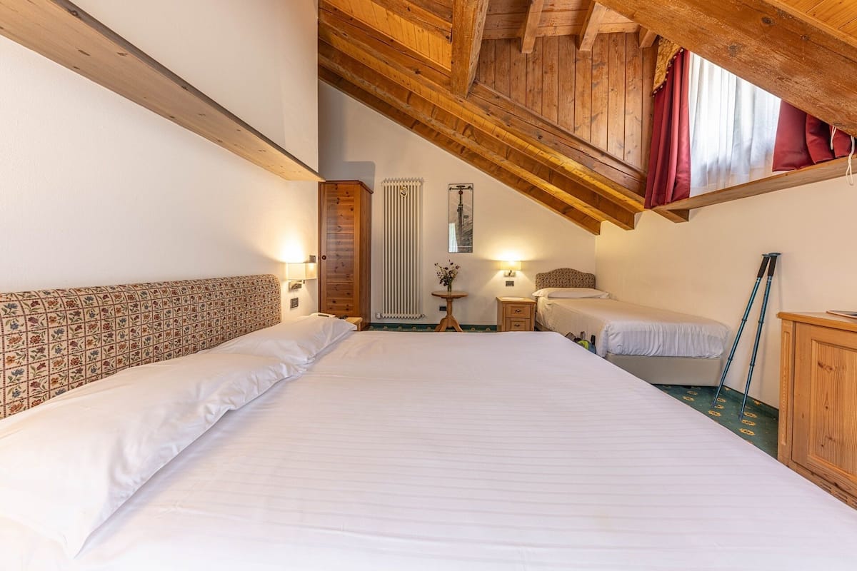 A bedroom with two beds, a wooden ceiling, a radiator, a small table with a vase, and a window with curtains.