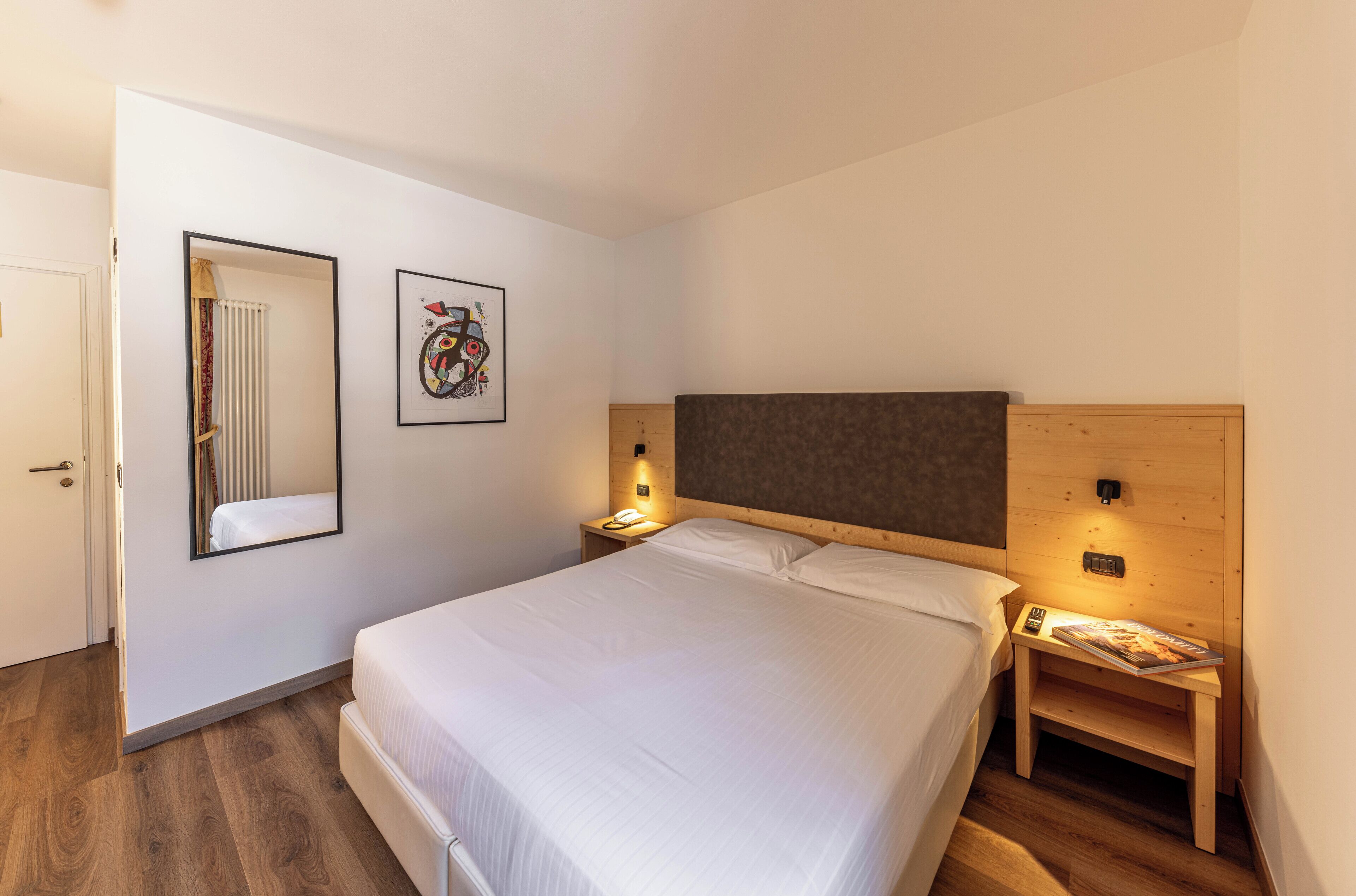 Ein Schlafzimmer mit einem großen Bett, einem Nachttisch mit Lampe, einem Wandspiegel und einem gerahmten Bild.