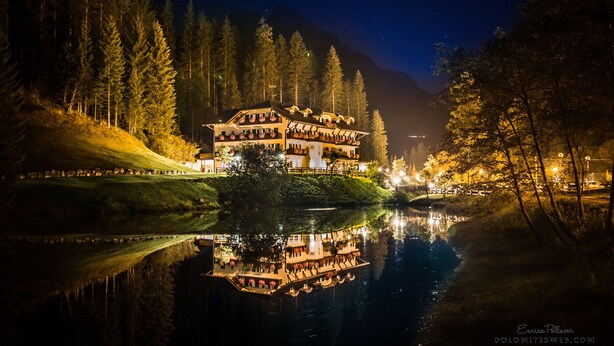 Un grand hôtel à plusieurs étages dont l’image se reflète dans l’eau, entouré d’arbres et avec une montagne en arrière-plan.