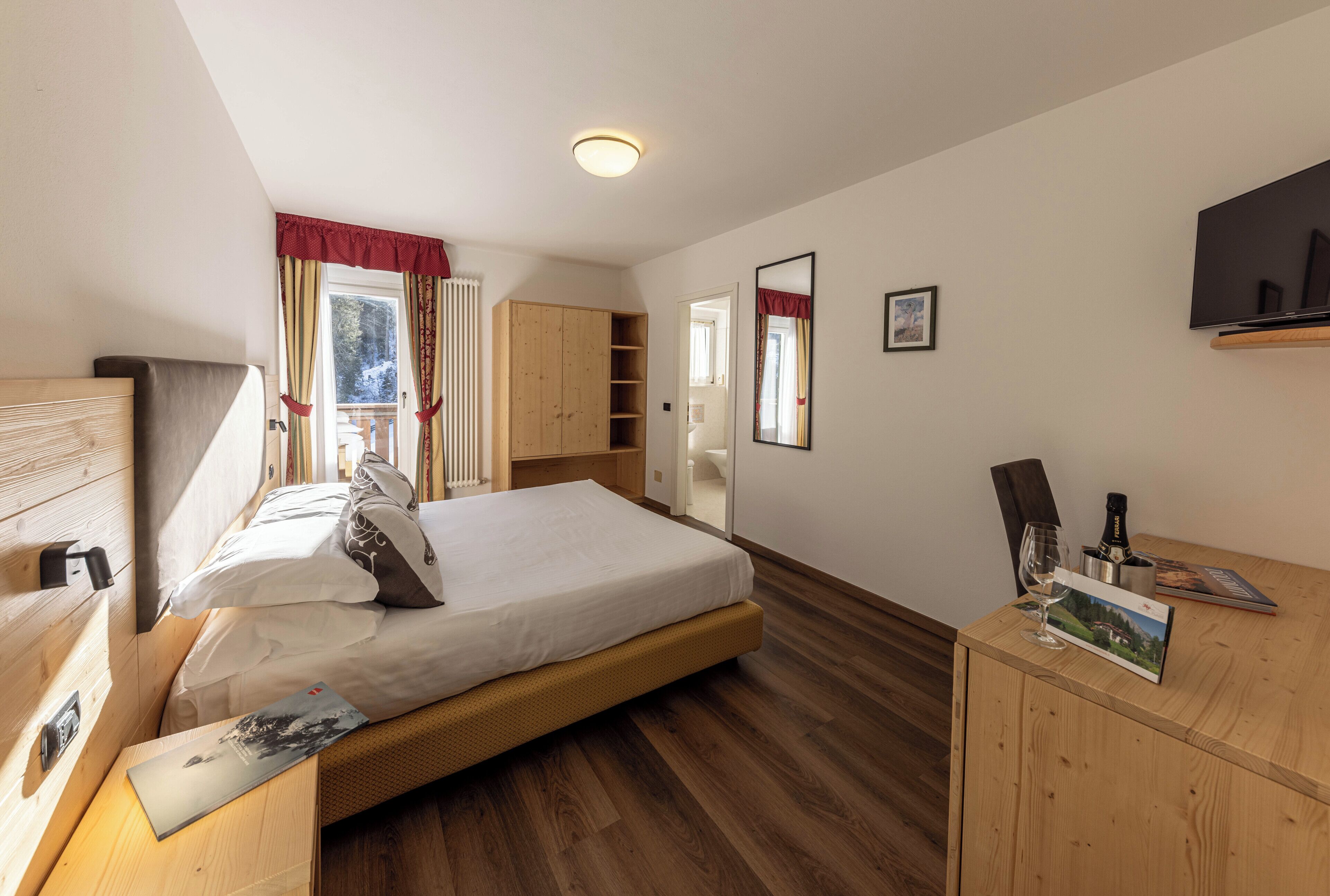 A bedroom with a bed, wooden wardrobe, a desk with a chair, a mirror, and a TV on the wall.