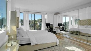 Un dormitorio moderno con una cama grande, un escritorio y una computadora. Hay ventanas amplias y vistas al exterior.