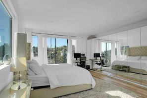 A modern bedroom with a large bed, a desk, and a computer. There are large windows and a view of the outdoors.