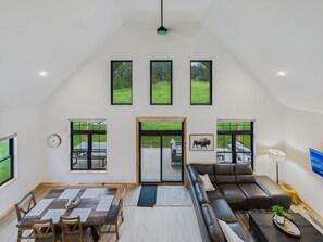 A spacious loft with a high ceiling, large windows, a dining area, a sofa, and a TV.