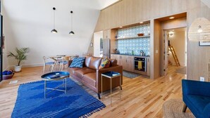 Un salon moderne avec un canapé, une table basse et un tapis bleu. On aperçoit une cuisine à l’arrière-plan, avec des meubles en bois et un pare-feu décoré.