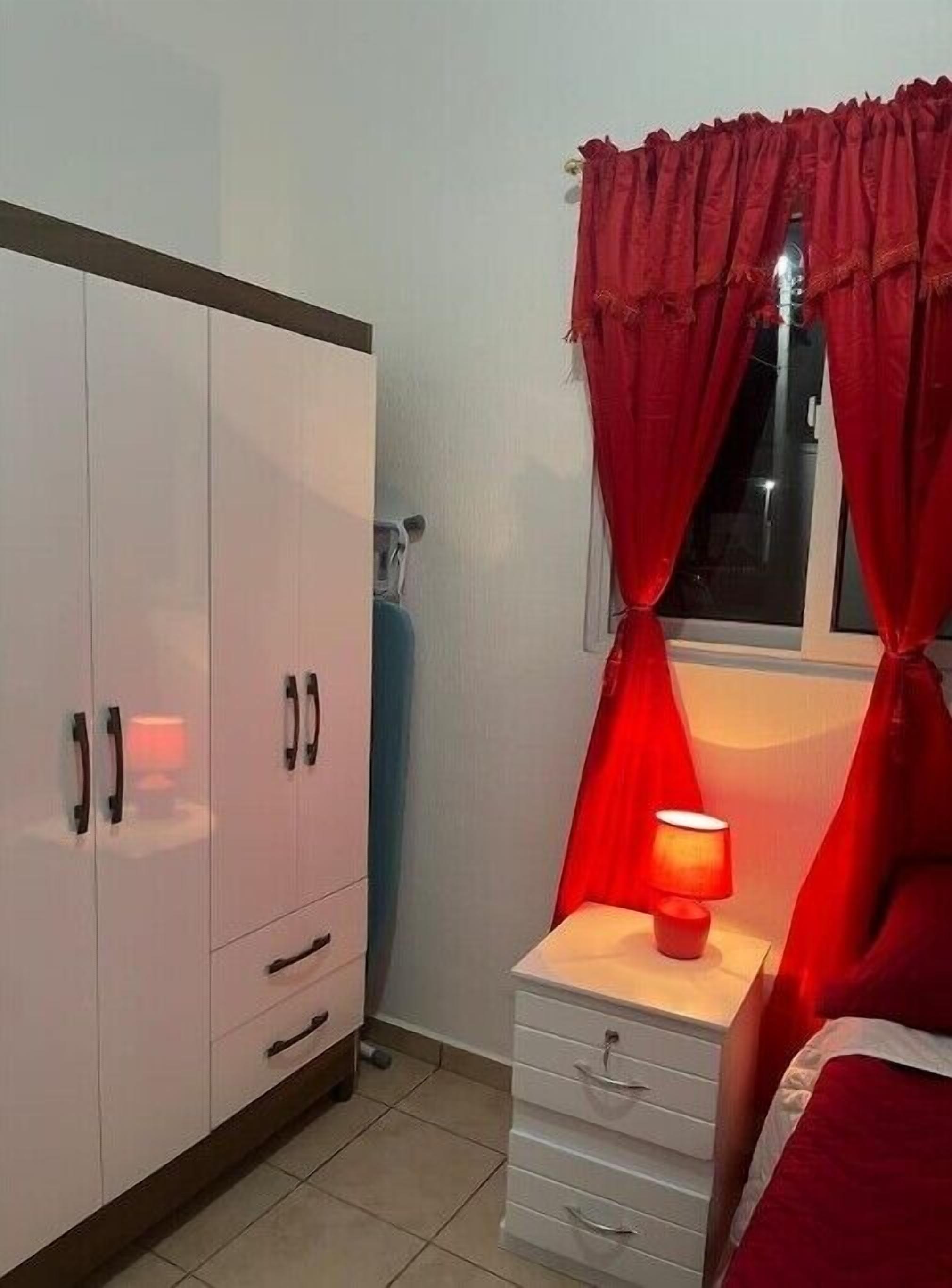 Quarto com guarda-roupa, mesa de cabeceira com abajur e janela com cortinas vermelhas.