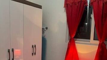 Quarto com guarda-roupa, mesa de cabeceira com abajur e janela com cortinas vermelhas.