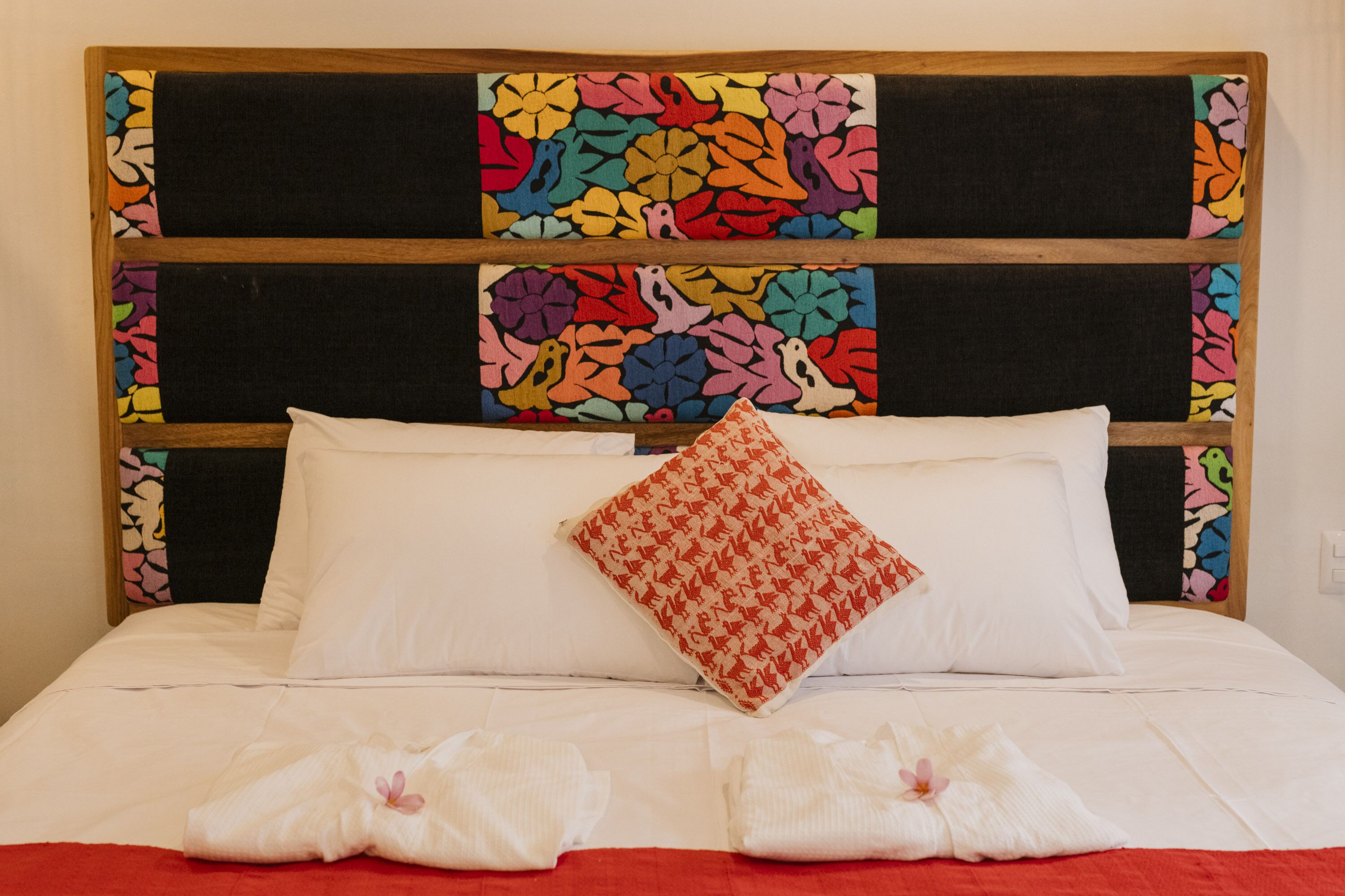Un lit bien fait, avec du linge de lit blanc, un coussin à motifs rouges et blancs, et une tête de lit colorée ornée de motifs floraux.