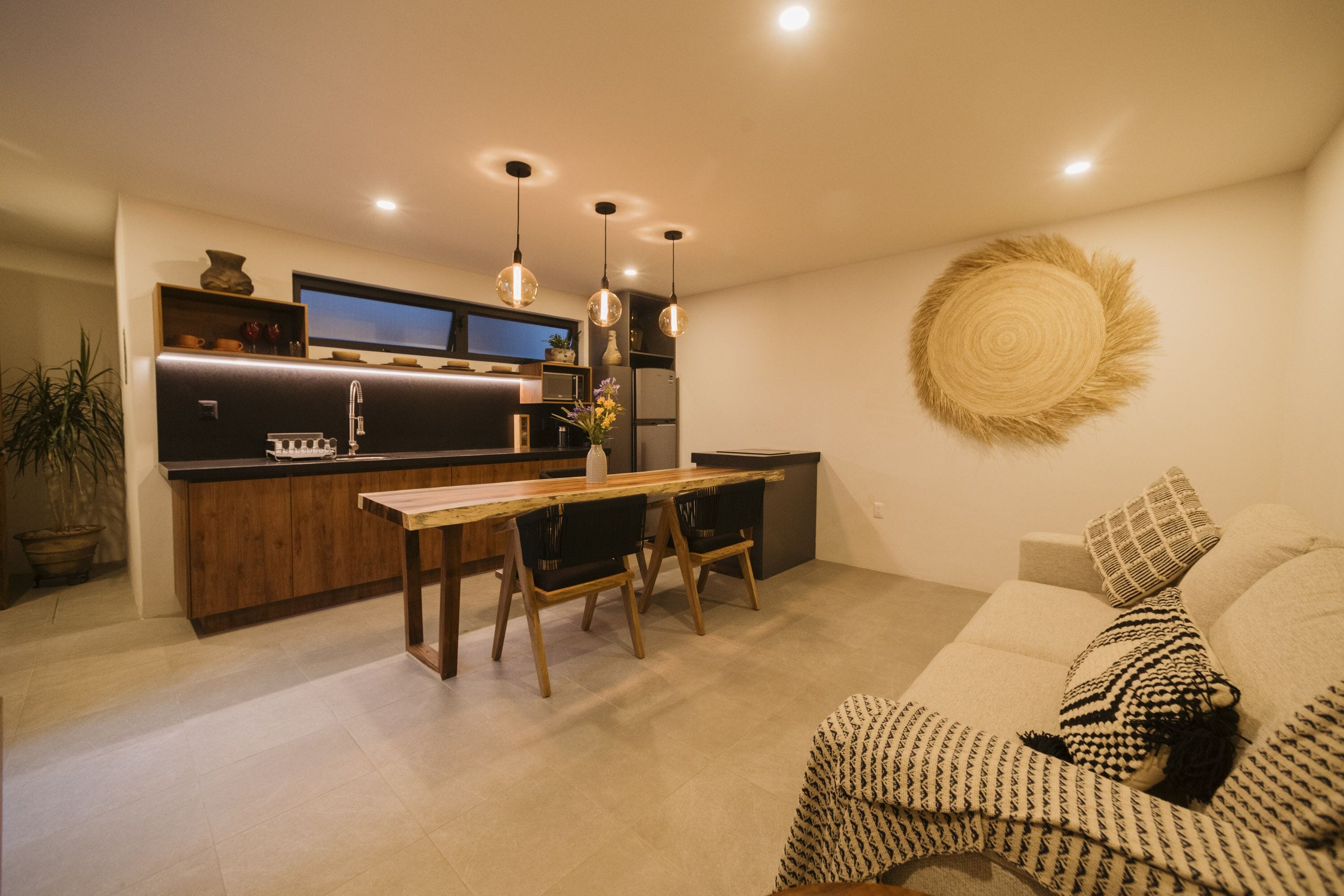 Eine moderne Küche mit Holzausstattung, einem Esstisch und Stühlen. Im Wohnbereich befindet sich ein großer Wandteppich und ein Blumentopf.