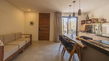 A modern living room with a sofa, a dining area with chairs, a kitchen with a counter and cabinets, and a sliding door leading to a balcony.