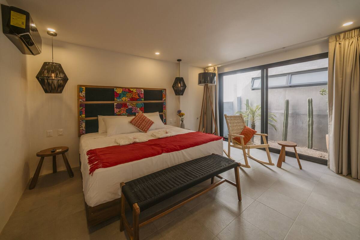 A modern bedroom with a large bed, a bench, a rocking chair, and a view of cacti through a large window.