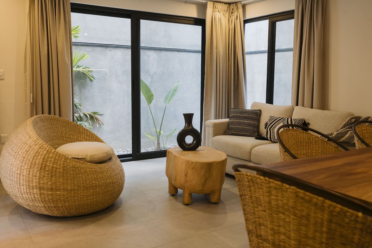 A modern living room with a large window, a sofa with patterned cushions, a round wicker chair, and a small wooden stool.