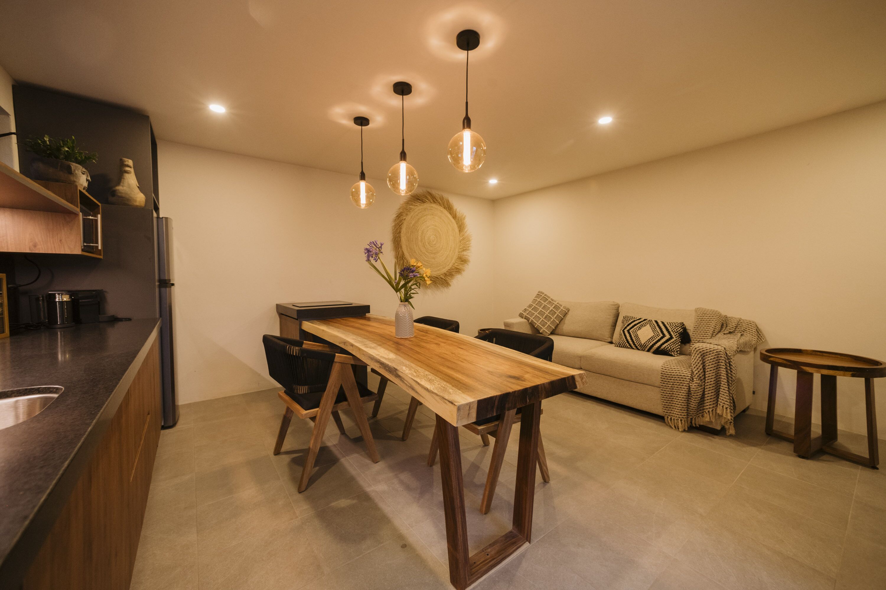 A modern kitchen with a wooden dining table, black chairs, and pendant lighting.