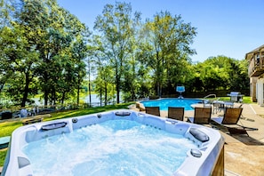 Un bain à remous entouré de meubles de jardin et une piscine, avec des arbres et un ciel dégagé en arrière-plan.