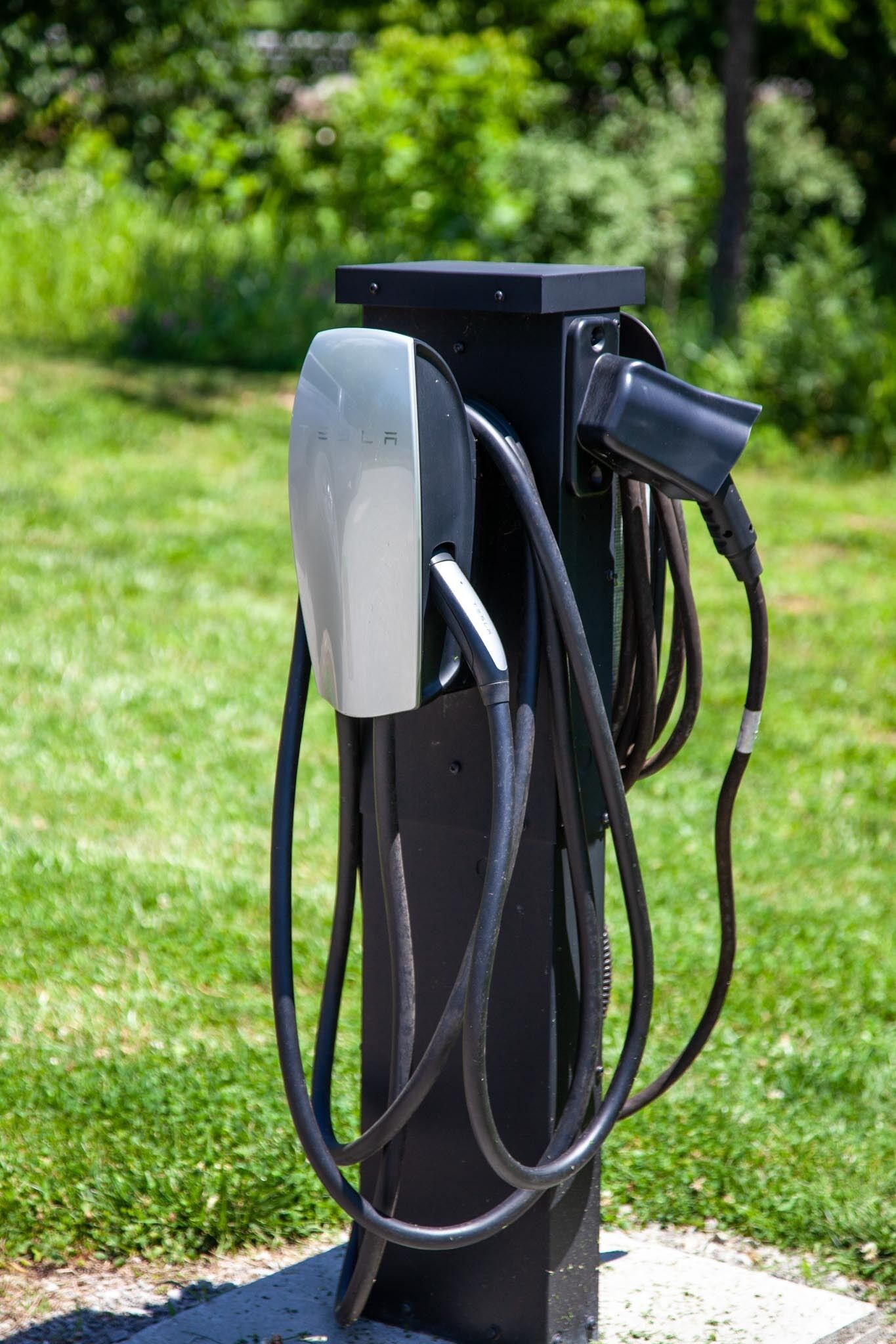 Een zwarte laadpaal voor elektrische auto's met meerdere kabels, geplaatst op een betonnen ondergrond, omringd door gras en bomen.